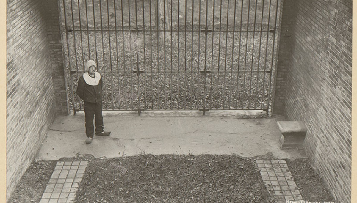 Maison d’arrêt de Fresnes : un détenu en cours de promenade individuelle - Photographie d’Henri Manuel, vers 1930 (Coll. Enap-Crhcp). Le détenu, en tenue pénale et sabots, a pu relever son capuchon une fois arrivé dans la cour de promenade individuelle. Il le baissera à nouveau en sortant de la cour et pour toute la durée de son déplacement vers sa cellule.